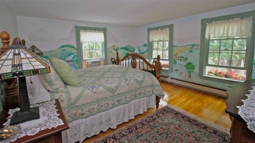 Cozy bedroom with a wooden bed, green accents, floral curtains, and scenic wall murals. Warm wooden floors and a rug.