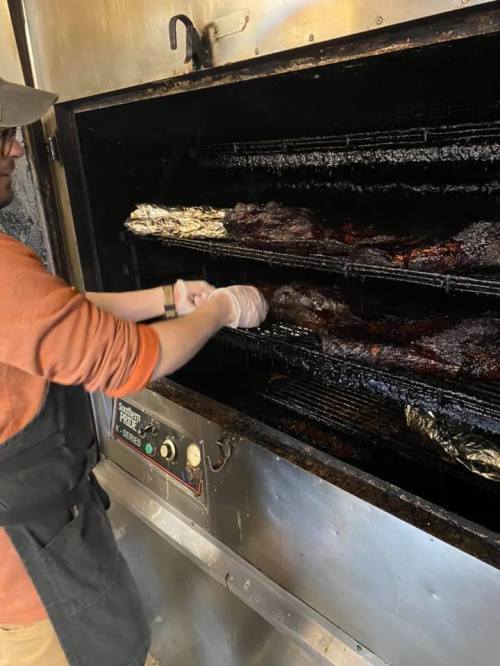 A person in an apron and gloves removes food from a large smoker, with smoke and foil-wrapped items visible inside.