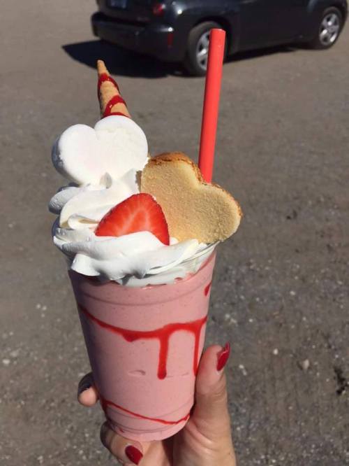 A hand holding a pink milkshake topped with whipped cream, a strawberry, heart-shaped cookies, and a straw.