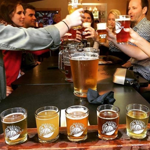 A group of people toasting with drinks at a bar, featuring a flight of beer samples in front of them.