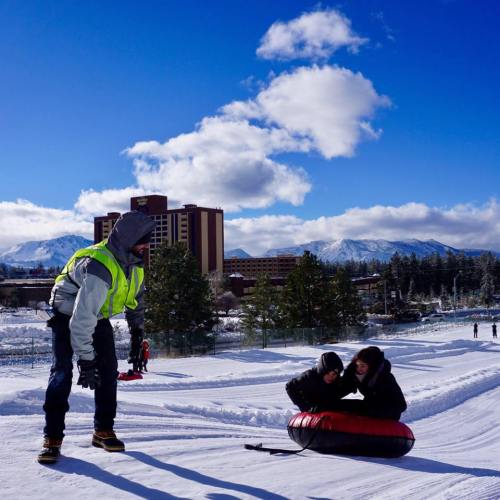 A person in a bright vest helps two people on a snow tubing hill with mountains and clouds in the background.