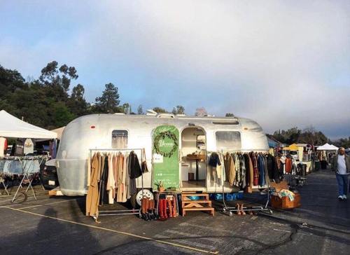 A vintage silver Airstream trailer serves as a clothing shop, with garments displayed outside at a market.