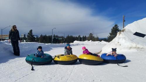Four children in colorful snow tubes sit on a snowy slope, with a woman standing nearby against a clear blue sky.