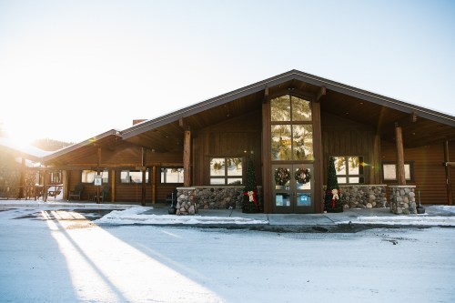 A modern wooden building with large windows, surrounded by snow, and decorated for the holidays. Sunlight shines in.