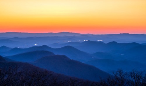 A serene mountain landscape at sunset, featuring layered hills and a gradient sky from orange to deep blue.