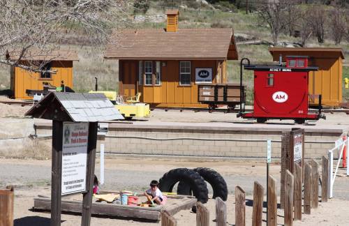 Two small buildings and a red train car in a sandy area, with children playing near a tire and wooden posts.