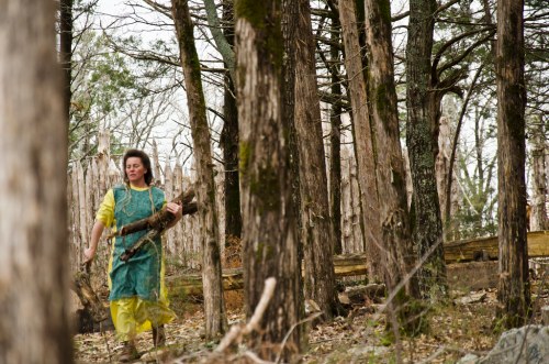 A woman in a green apron and yellow dress walks through a forest, carrying a bundle of sticks.