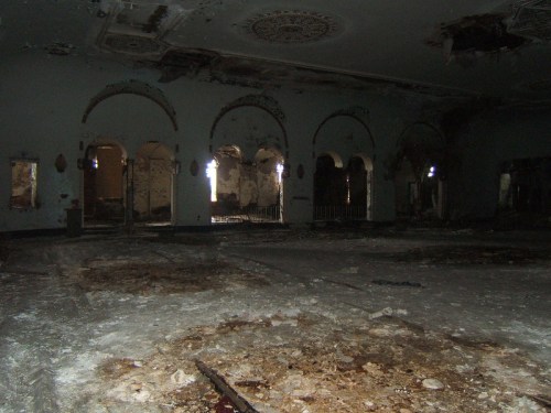 Abandoned room with crumbling walls, arched doorways, and debris scattered on the floor, illuminated by faint light.