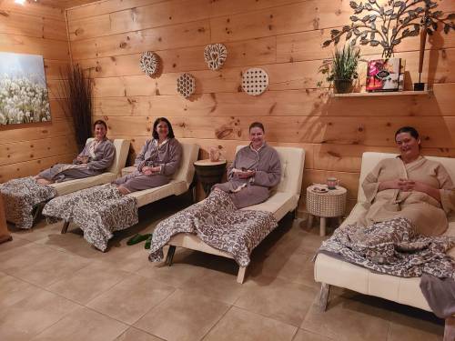 Four women in robes relax on lounge chairs in a cozy spa setting with wooden walls and soft lighting.