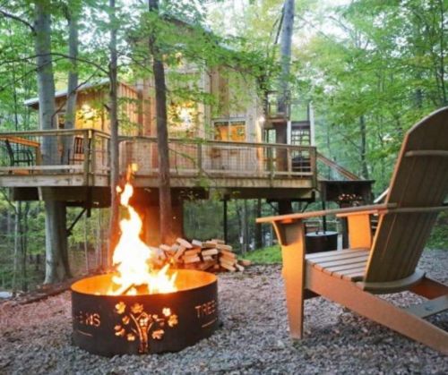 A cozy treehouse surrounded by trees, with a fire pit and an Adirondack chair in the foreground.