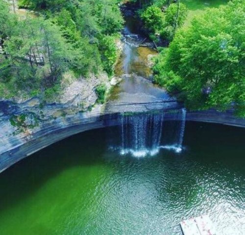 Aerial view of a circular waterfall flowing into a green lake, surrounded by lush trees and a rocky cliff.