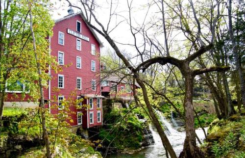 A red historic mill building beside a flowing stream, surrounded by lush green trees in a serene natural setting.