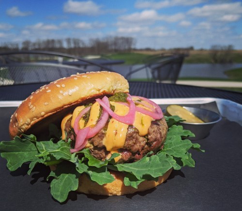 A juicy burger with lettuce, pickled onions, and cheese, served on a sesame seed bun with a side of pickles.