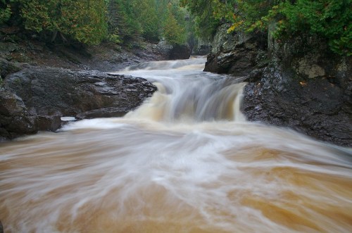 A flowing river cascades over rocks, surrounded by lush green trees in a serene natural setting.