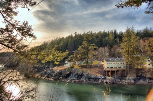 A serene riverside scene featuring a house on stilts, surrounded by trees and rocky shores under a cloudy sky.