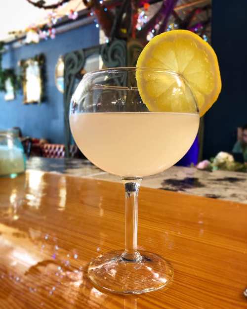 A cocktail in a coupe glass garnished with a lemon slice, sitting on a wooden bar top.