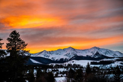 A stunning sunset over snow-capped mountains, with vibrant orange and pink hues in the sky and silhouetted trees in the foreground.