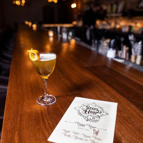 A cocktail with a lemon twist sits on a wooden bar next to a menu, with soft lighting in the background.
