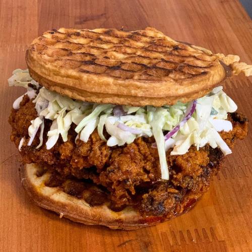 A crispy fried chicken sandwich with lettuce and sauce, served between two waffle halves on a wooden surface.