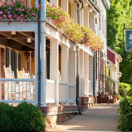 Charming porch with rocking chairs and vibrant flower boxes, leading to a quaint inn in a sunny, tree-lined setting.