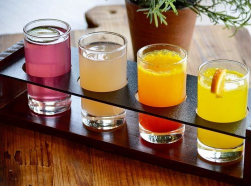 A wooden tray holds four colorful drinks: pink, light yellow, orange, and yellow, with a plant in the background.