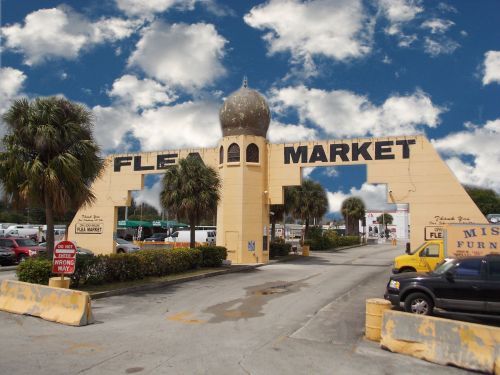 Entrance to a vibrant flea market with a large sign, palm trees, and a blue sky with fluffy clouds.