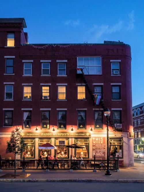 A lively pub, Paddy Murphy's, illuminated at dusk, with outdoor seating and a rainbow flag displayed.