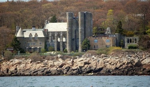A stone mansion with towers sits on a rocky shoreline, surrounded by trees and hills in the background.