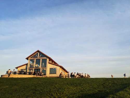 A wooden building on a grassy hill with people sitting outside, enjoying the view under a clear blue sky.