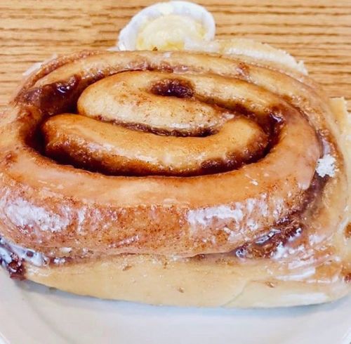 A close-up of a cinnamon roll with a glossy glaze, served with a small cup of butter on a white plate.