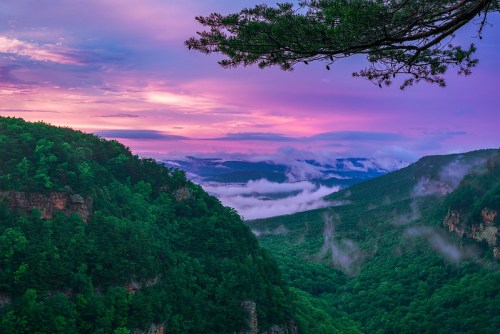 A vibrant sunset over misty mountains, with lush green valleys and a colorful sky filled with purple and pink hues.