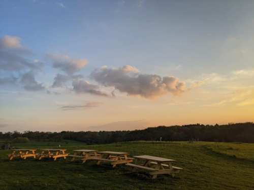 A serene landscape featuring picnic tables on green grass under a colorful sunset sky with scattered clouds.