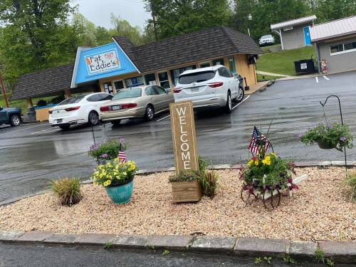 A welcoming sign and flower pots in front of a restaurant named "Fat Eddie's," with parked cars and a rainy atmosphere.