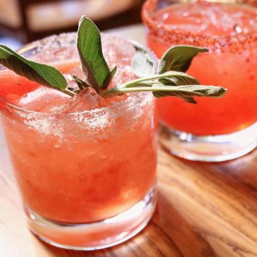 Two cocktails with ice, garnished with fresh sage leaves, served in clear glasses on a wooden table.