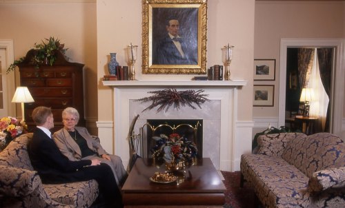 A cozy living room with two people sitting on a sofa, a fireplace, and a large portrait on the wall.
