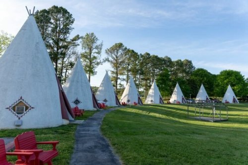 A row of white teepees with red accents, surrounded by green grass and trees, with red chairs in front.