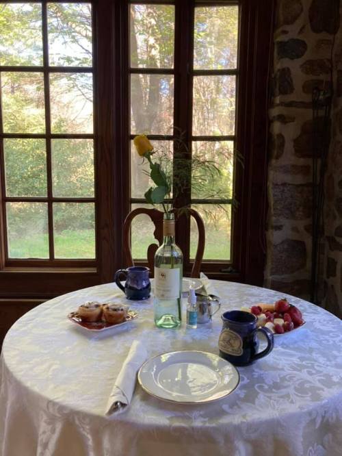 A cozy table set with a white tablecloth, wine, a rose, pastries, and fruit, beside large windows with a garden view.