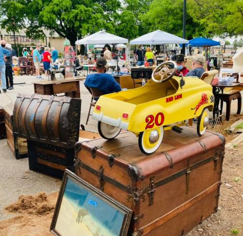A yellow toy car sits on a stack of vintage trunks at an outdoor market, with people browsing in the background.
