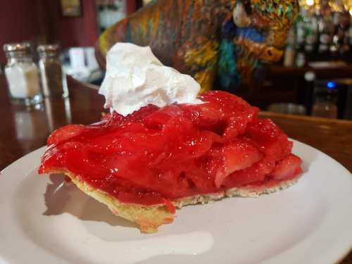 A slice of strawberry pie topped with whipped cream on a white plate, with a colorful background.