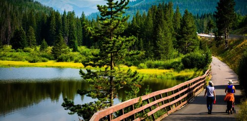 Two people walk along a path by a serene lake, surrounded by lush greenery and mountains in the background.