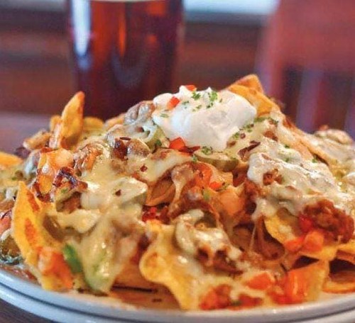 A plate of loaded nachos topped with cheese, vegetables, and sour cream, with a glass of beer in the background.