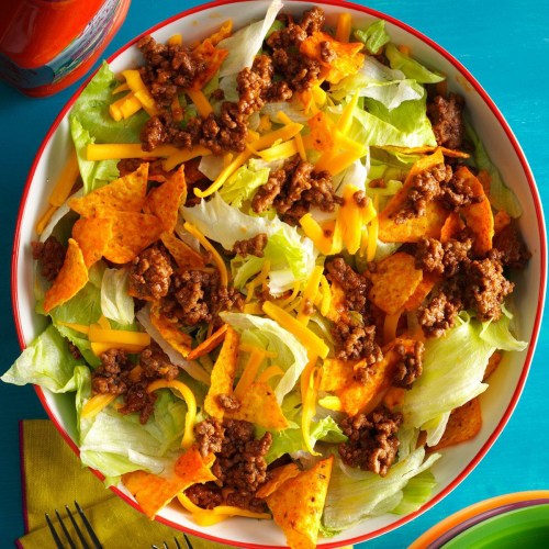 A colorful salad with lettuce, ground beef, cheese, and tortilla chips in a large bowl on a bright blue table.