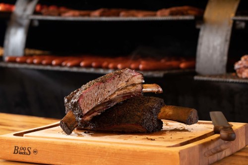 A close-up of a smoked beef rib on a wooden cutting board, with a blurred background of sausages on a grill.