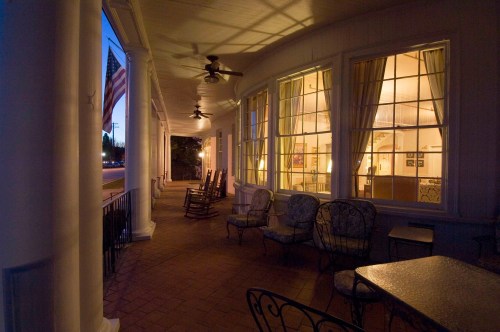 A cozy porch with rocking chairs and a table, illuminated by warm light from the windows, set against a twilight sky.