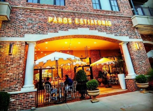 Exterior of Paros Estiatorio, featuring outdoor seating under umbrellas and a brick facade. Warm evening ambiance.