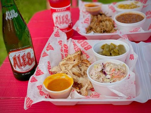 A takeout meal featuring pulled chicken, coleslaw, beans, pickles, and Ale-8 soft drink on a red table.