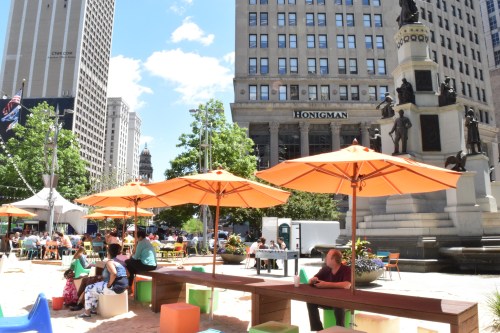 A vibrant outdoor space with orange umbrellas, people dining, and a monument in a city setting on a sunny day.