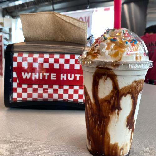 A milkshake topped with whipped cream and sprinkles, with a red straw, in front of a checkered "White Hut" sign.