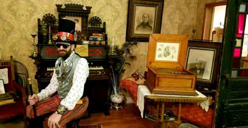A man in a top hat and sunglasses sits in a vintage room with an antique phonograph and framed portraits.
