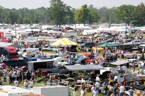 A bustling outdoor market with numerous tents, vehicles, and crowds of people browsing various items.